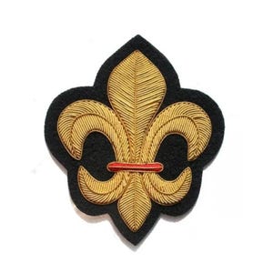 May include: A gold embroidered fleur-de-lis badge on a black felt backing. The design features a red horizontal band. This emblem is a traditional symbol, commonly seen in heraldry and decorative applications.