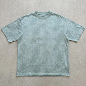 May include: A short-sleeved, crew neck t-shirt in a muted, seafoam green colour. The fabric has a textured, mottled appearance, giving it a vintage or worn look. The shirt is a basic, casual style.