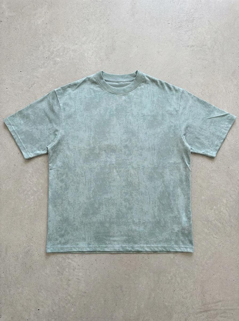 May include: A short-sleeved, crew neck t-shirt in a muted, seafoam green colour. The fabric has a textured, mottled appearance, giving it a vintage or worn look. The shirt is a basic, casual style.