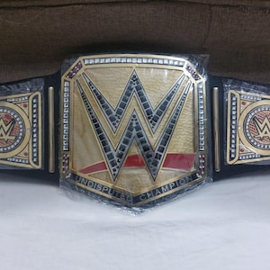 Puede incluir: Un cinturón de campeonato de lucha libre dorado y negro con el logotipo de la WWE. El cinturón tiene una placa central grande con las letras "W" y "W" en dorado, y las palabras "Undisputed Champion". Las placas laterales también presentan el logotipo de la WWE.
