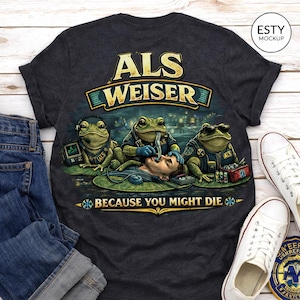 May include: Dark gray t-shirt with a graphic featuring three frogs in medical attire attending to a patient. The text "ALS WEISER" is above the image, and "BECAUSE YOU MIGHT DIE" is below.