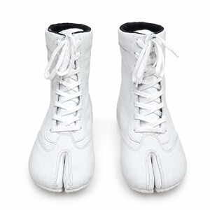 May include: A pair of white high-top boots featuring a split-toe design, white laces, and a black interior collar lining. The boots have a smooth finish and a lace-up front closure that extends from the ankle down to the top of the foot area.