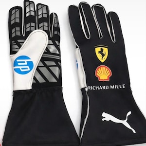 May include: A pair of black racing gloves featuring white accents and logos. The gloves display the HP, Ferrari, Shell, Richard Mille, and Puma branding. The palm side includes a grey geometric pattern, while the wrist area is solid black.