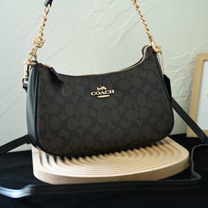 May include: A brown and black Coach shoulder bag with a gold chain strap and zipper. The bag has a repeating pattern and a gold Coach logo. The bag is on a wooden tray.