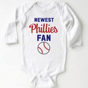 May include: White long-sleeved baby bodysuit with the text "NEWEST Phillies FAN" in red and blue. A baseball graphic is below the text. The bodysuit has a snap closure at the bottom.