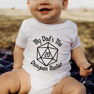 May include: A white baby onesie with the text "My Dad's The Dungeon Master" in black, featuring a 20-sided die graphic. The baby is sitting on a plaid blanket outdoors.
