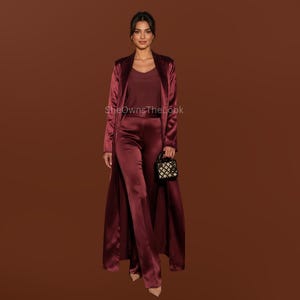 May include: A deep burgundy satin ensemble consisting of a long-line open duster coat, a sleeveless v-neck top, and matching wide-leg trousers. The outfit is paired with a small black structured handbag featuring a gold-tone geometric pattern.