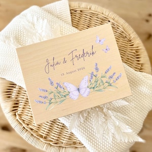 Könnte beinhalten: Ein Holzschild mit den Namen "Julia & Frederik" und dem Datum "13. August 2026" in lila Schrift. Das Schild ist mit Lavendelzweigen und Schmetterlingen in Lila- und Weißtönen verziert. Das Schild liegt auf einer weißen Decke.