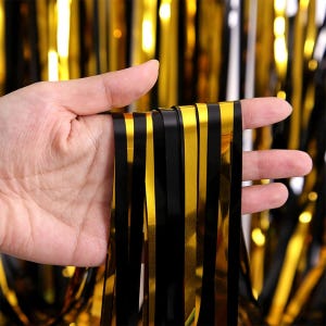 Puede incluir: Cortina de flecos metálicos negros y dorados. Las brillantes tiras verticales alternan negro y dorado, creando un efecto decorativo. El fondo muestra más flecos, ideal para fiestas y celebraciones.