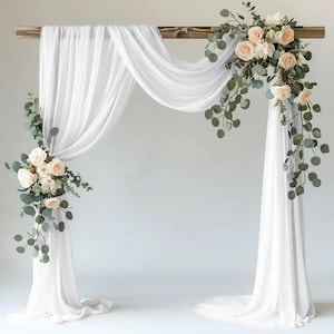 Könnte beinhalten: Eine Hochzeitsbogen aus Holz, drapiert mit fließendem weißem Stoff und geschmückt mit floralen Arrangements aus cremefarbenen Rosen und grünem Laub. Der Bogen steht vor einem schlichten weißen Hintergrund.