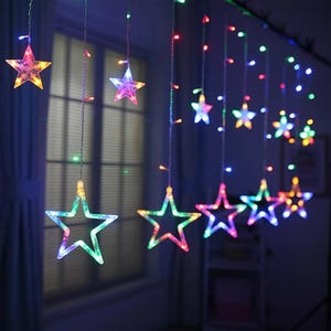 May include: A string of colorful star-shaped lights. The stars are outlined in various colors, including green, blue, pink, and yellow. The lights are hanging in front of a window, creating a festive and decorative display.