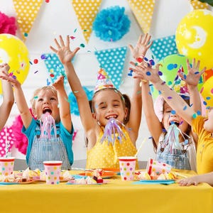 May include: A group of children celebrating a party, with confetti and party hats. The image shows kids with their arms raised, confetti falling, and party hats. Yellow balloons and a yellow tablecloth add to the festive atmosphere. Polka dot cups and plates are on the table.
