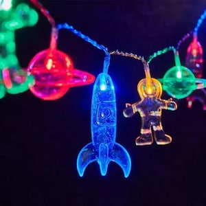 Może przedstawiać: Szereg kolorowych świateł LED z kształtami o tematyce kosmicznej, w tym niebieska rakieta, żółty astronauta oraz czerwone i zielone planety. Ciemne tło.