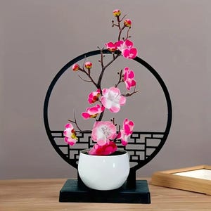 Puede incluir: Arreglo floral decorativo con flores rosas y blancas. Las flores están en una maceta de cerámica blanca, frente a una pantalla decorativa negra y circular. La base es un cuadrado negro. Un elemento decorativo para el hogar.