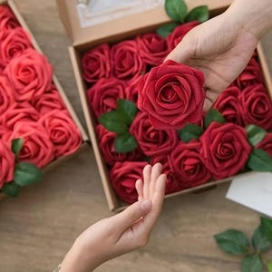 May include: A person holds a single, deep red artificial rose above a box filled with similar blooms. The roses have green leaves and are arranged in a cardboard box. The roses are a vibrant, rich red colour.