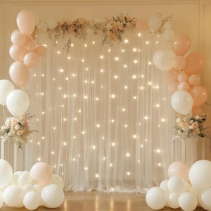 Pode incluir: Um cenário decorativo com uma cortina branca transparente iluminada por pequenas luzes. Aglomerados de balões brancos e cor de rosa são dispostos em ambos os lados, com arranjos florais e uma grinalda de flores no topo.