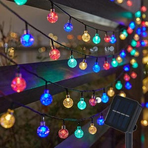 Puede incluir: Una cadena de luces LED solares con pequeñas esferas translúcidas y texturizadas en varios colores, incluyendo azul, rojo, amarillo y verde. Las luces están conectadas por un cable negro e incluyen una unidad de panel solar negra.