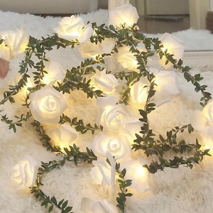 Puede incluir: Guirnalda de luces LED con forma de rosas blancas y detalles de hojas verdes. Las luces emiten un brillo cálido, ideal para decoración. La guirnalda mide aproximadamente 200 cm.