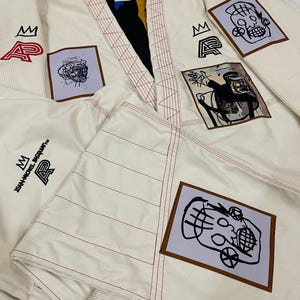 May include: Off-white martial arts uniform with red stitching and patches featuring abstract art and text. The uniform has a black and yellow lining and the text "JEAN-MICHEL BASQUIAT" is visible.
