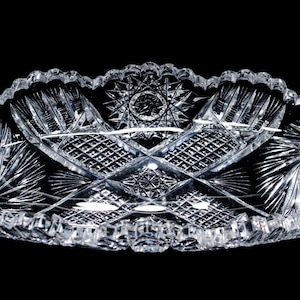 May include: An oval clear glass serving dish featuring intricate cut-glass patterns including starburst motifs, cross-hatching, and fan-shaped details. The edges of the dish are scalloped with a serrated design, creating a reflective surface.