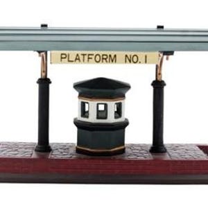Puede incluir: Un modelo en miniatura de andén de estación de tren con una base de adoquines de color rojo oscuro, dos pilares de soporte negros y un techo verde. Un letrero colgado del techo dice PLATFORM NO. 1. Una pequeña caseta de señales hexagonal.