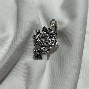 Può includere: Un ciondolo a forma di drago color argento con dettagli intricati, tra cui squame, ali e una coda arricciata. Il ciondolo è sospeso da un piccolo anello, suggerendo che sia progettato per essere indossato come un ciondolo da collana. Sfondo bianco.