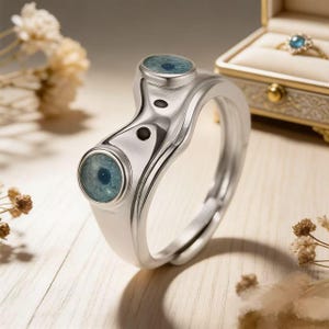 May include: A silver ring with an eye design. The ring features two round, light blue eye-shaped stones and two small black dots. The ring is adjustable and sits on a light wooden surface. A jewelry box with a gold ring is in the background.