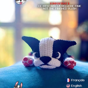 Peut inclure: Peluche crochetée représentant un Boston Terrier noir et blanc, avec des coussinets roses. Le texte indique "Suki - Boston Terrier" et "CE N'EST PAS LA PELUCHE FINIE". Le jouet est posé sur une surface bleue.