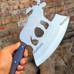 May include: A large, silver-colored axe with a moose silhouette design. The axe has a blue and brown handle. The blade has three cut-out shapes. The text "Northridgeblads" is visible on the blade. The axe is held in a person's hand.