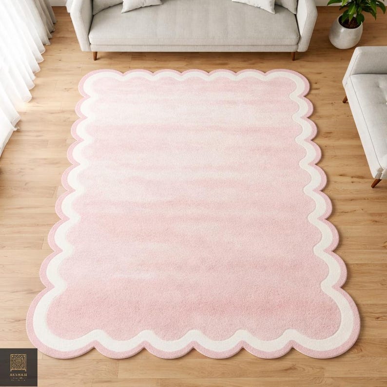 May include: A large, rectangular pink rug with a scalloped edge. The rug has a watercolor-like effect, with a gradient from light to darker pink. A white border and a light pink outline the scalloped edge.