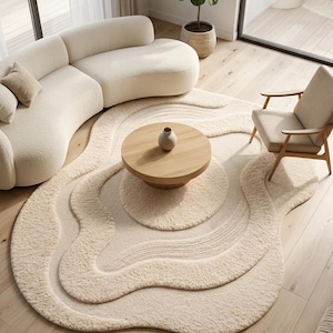 Può includere: Un tappeto color bianco sporco con una forma organica, ondulata e stratificata, caratterizzato da una texture a pelo lungo. Il tappeto presenta motivi concentrici, rialzati e increspati che imitano le formazioni di sabbia, creando una superficie.