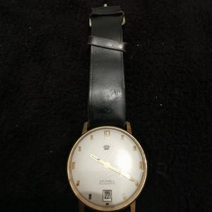 Könnte beinhalten: Eine vintage goldfarbene Armbanduhr mit weißem Zifferblatt, schwarzem Lederarmband und dem Datum 22. Das Zifferblatt der Uhr hat goldene Zeiger und ein Kronen-Emblem. Die Uhr ist mit "DRUKELS AUTOMATIC" beschriftet.