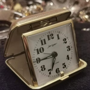 May include: A gold-toned Jerger alarm clock with a white face and black numerals. The clock is in a rectangular case with a hinged lid. The clock face displays the word "alarm".