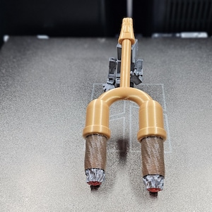 May include: A 3D-printed bicycle fork in gold and brown. The fork has a curved design with two lower legs and a top section. The lower legs are brown with a textured pattern and red accents at the bottom.