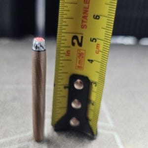 May include: A small, cylindrical, metallic bronze-colored object with a silver-toned, ridged cap at the top. It is positioned next to a yellow measuring tape, which indicates that the object measures approximately 1.75 inches in total height.