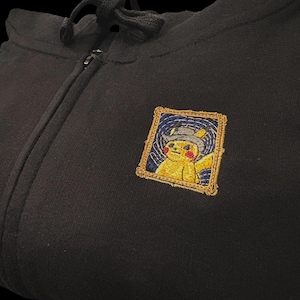 May include: Black zip-up hoodie with a small embroidered patch. The patch features a yellow Pikachu character wearing a hat, framed in gold. The hoodie has a drawstring hood and a full-length zipper.