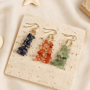 Könnte beinhalten: Drei Paar Ohrhänger mit goldfarbenen Haken und Strängen aus unregelmäßigen, getrommelten Edelstein-Splittern. Das Set enthält ein Paar aus tiefblauem Lapislazuli, ein Paar aus durchscheinendem orangefarbenem Karneol und ein Paar aus hellgrünem Aventurin.