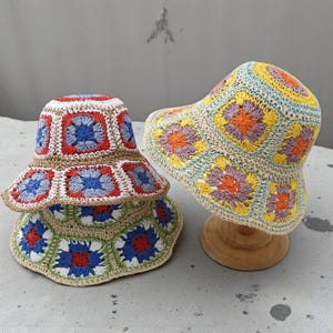 Op de afbeelding: Twee kleurrijke gehaakte bucket hats. Eén hoed heeft rode, blauwe en witte vierkanten, de andere gele, oranje en paarse vierkanten. Beide hoeden hebben een brede rand en worden op houten standaards gepresenteerd.