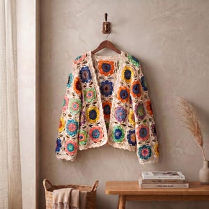 May include: A long-sleeved cardigan made of cream-colored yarn featuring a multi-colored crochet granny square pattern. The squares include vibrant shades of orange, blue, yellow, pink, and green, arranged in a repeating floral-inspired design.