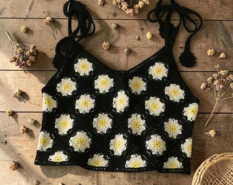 Puede incluir: Top de crochet negro con un patrón de margaritas. Las margaritas son blancas con centros amarillos. Las correas son negras y se atan a los hombros. El top es de estilo cuello en V.