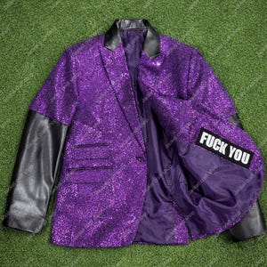 Op de afbeelding: Een paarse blazer met pailletten en zwarte leren mouwen en kraag. De binnenvoering heeft een patch met de woorden "FUCK YOU" in witte blokletters. De jas is open en onthult de binnenvoering.