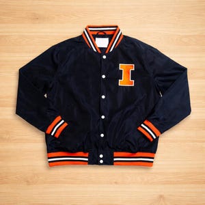 May include: Navy blue varsity jacket with orange and white striped details on the collar, cuffs, and hem. It has a large orange "I" patch on the chest. The jacket fastens with white press studs.