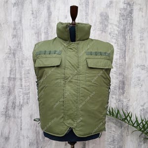 May include: Olive green vest with a high collar and two front pockets. The vest has a quilted design and reinforced shoulder areas. The vest is displayed on a mannequin. The text "Jacket craft shop" is visible.