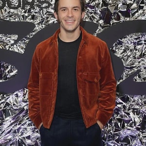 May include: A rust-colored corduroy jacket with two front pockets and a button closure. The jacket is worn over a black crew neck shirt and paired with dark blue pants. The background is a shiny silver and black surface.