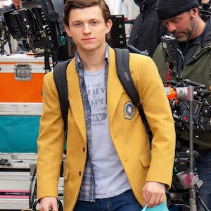 Op de afbeelding: Een mosterdgele blazer met een embleem op de linker borstzak. De blazer wordt gedragen over een grijs t-shirt met de tekst "IF YOU LIE MINE" en een geruit overhemd. Een zwarte rugzak wordt over de schouders gedragen.