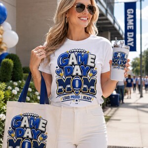 May include: A white t-shirt and a matching canvas tote bag, both featuring a blue and gold graphic with the text "GAME DAY MOM" and "LOUD PROUD & READY" alongside a football illustration. A silver tumbler displays the same matching design.