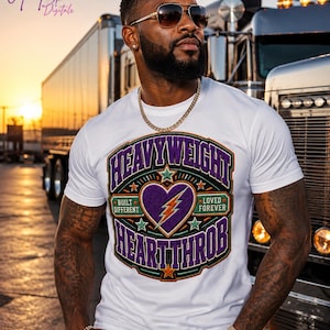 May include: A white short-sleeved t-shirt featuring a large, colourful graphic on the chest. The design includes the text HEAVYWEIGHT HEARTTHROB, BUILT DIFFERENT, and LOVED FOREVER, surrounding a purple heart with a yellow lightning bolt.