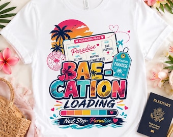 May include: A white t-shirt featuring a colorful graphic print of a boarding pass, a luggage tag, and the text "BAE CATION LOADING". The design includes palm trees, a sunset, a loading bar, and the phrase "Next Stop: Paradise" in a vibrant, tropical style.