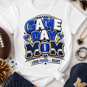 May include: A white short-sleeved t-shirt featuring a graphic design with the text "GAME DAY MOM" in blue and white block letters. A blue football replaces the letter O in MOM. The text "LOUD PROUD & READY" appears below in blue.