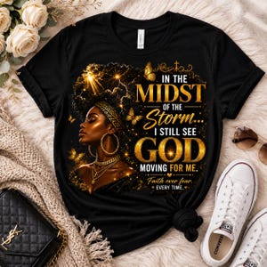 K&ouml;nnte beinhalten: Schwarzes T-Shirt mit einer goldenen Illustration einer Frau mit Goldschmuck und Schmetterlingen. Der Text lautet: "IN THE MIDST OF THE STORM... I STILL SEE GOD MOVING FOR ME. Faith over fear. EVERY TIME."